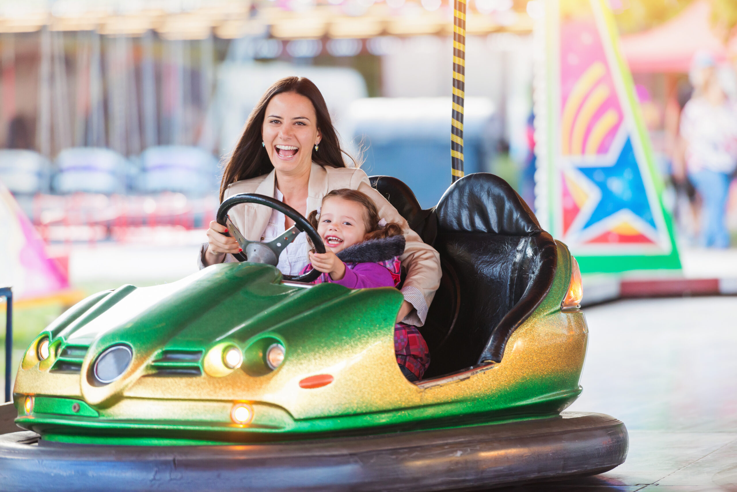 Cute little girl with her mother having fun at fun fair, driving a bumper car, amusement park