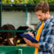 rancher in plaid shirt writing on clipboard near cowshed on farm