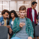 Serious young man sitting and using cell phone while his friends standing and talking