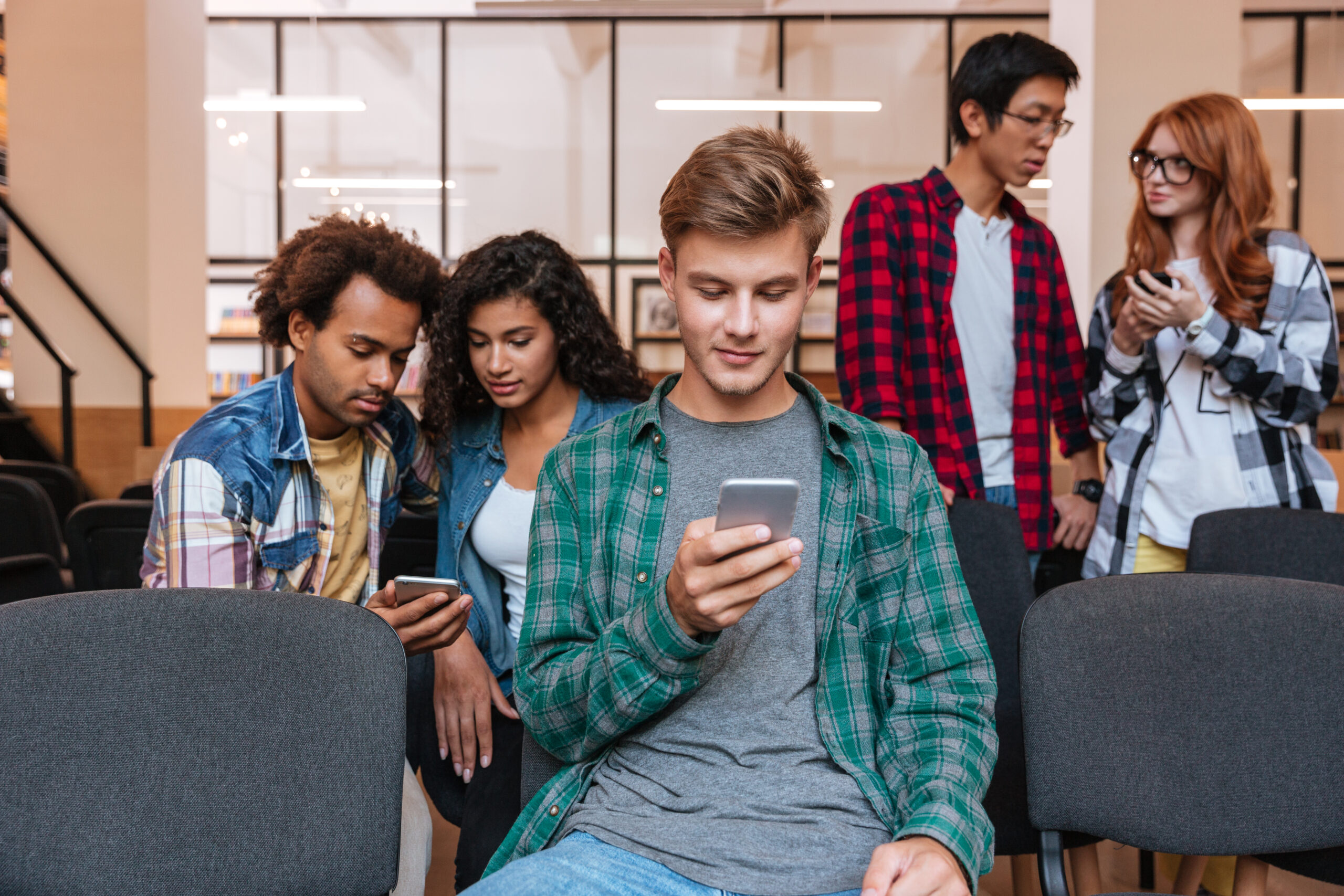 Serious young man sitting and using cell phone while his friends standing and talking