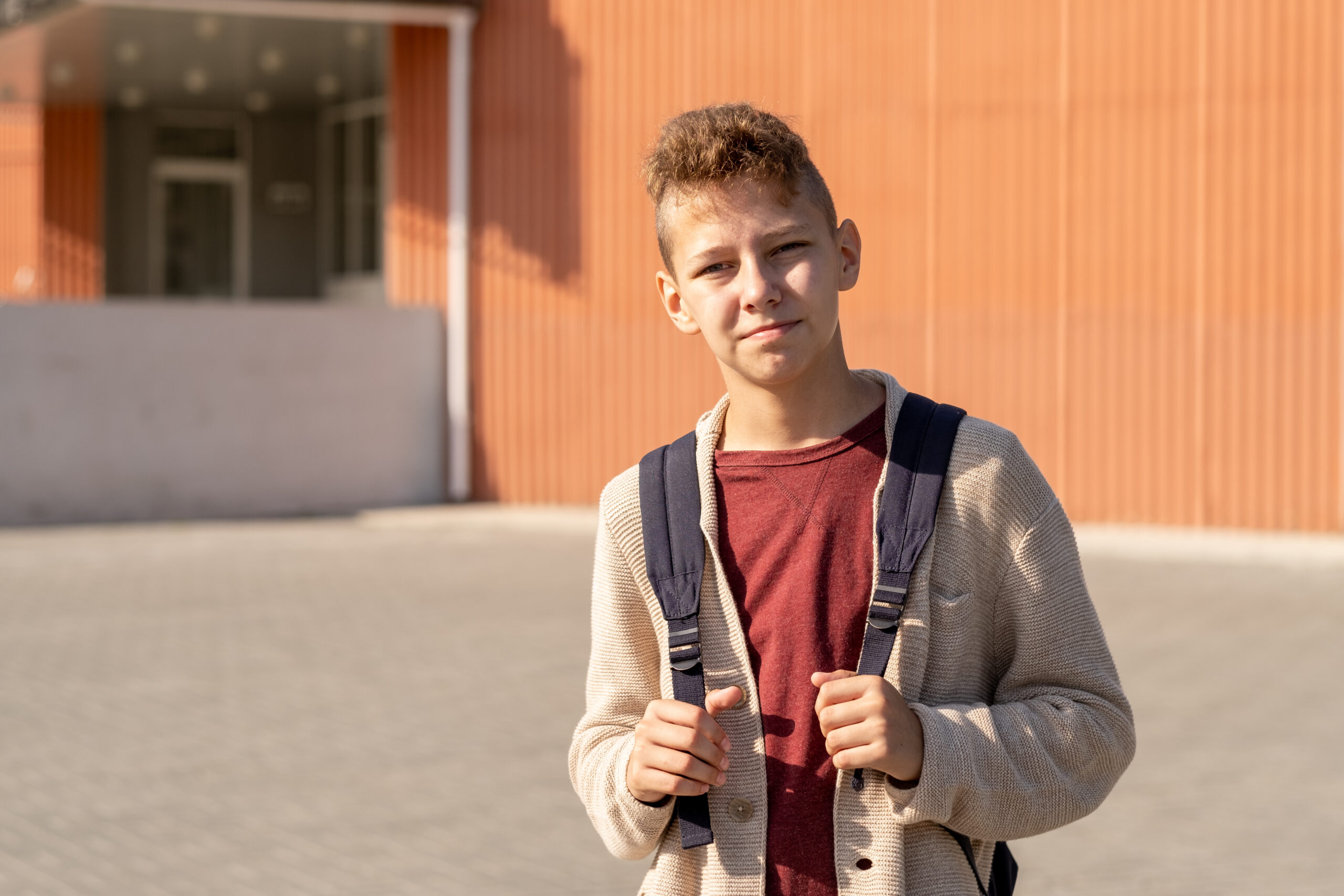 Adolescent boy with backpack standing against school building in front of camera