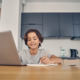 Smiling Caucasian boy spending time inside in the kitchen and using his computer while writing with pencil