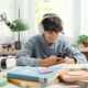 Adolescent student sitting at desk at home and doing homework, he is connecting with his smartphone and smiling