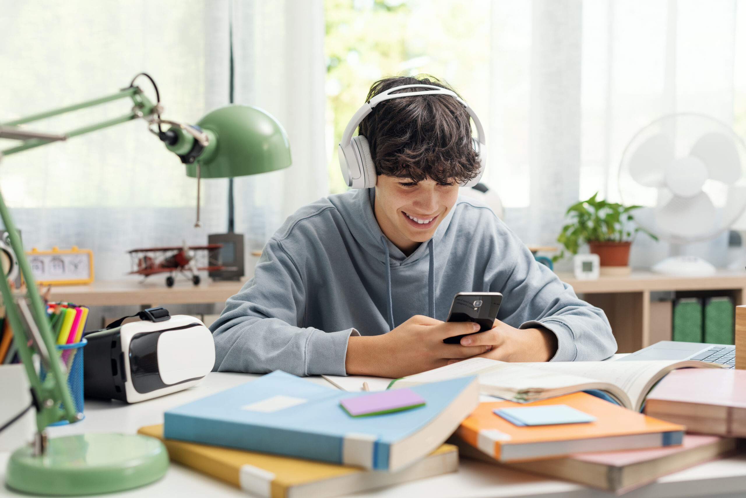 Adolescent student sitting at desk at home and doing homework, he is connecting with his smartphone and smiling