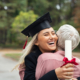 Enthusiastic graduated daughter holding degree embracing mother in campus. Young female student graduate hugging her mother at graduation ceremony. Excited college student with the graduation gown and hat holding diploma and hug the parent at campus.