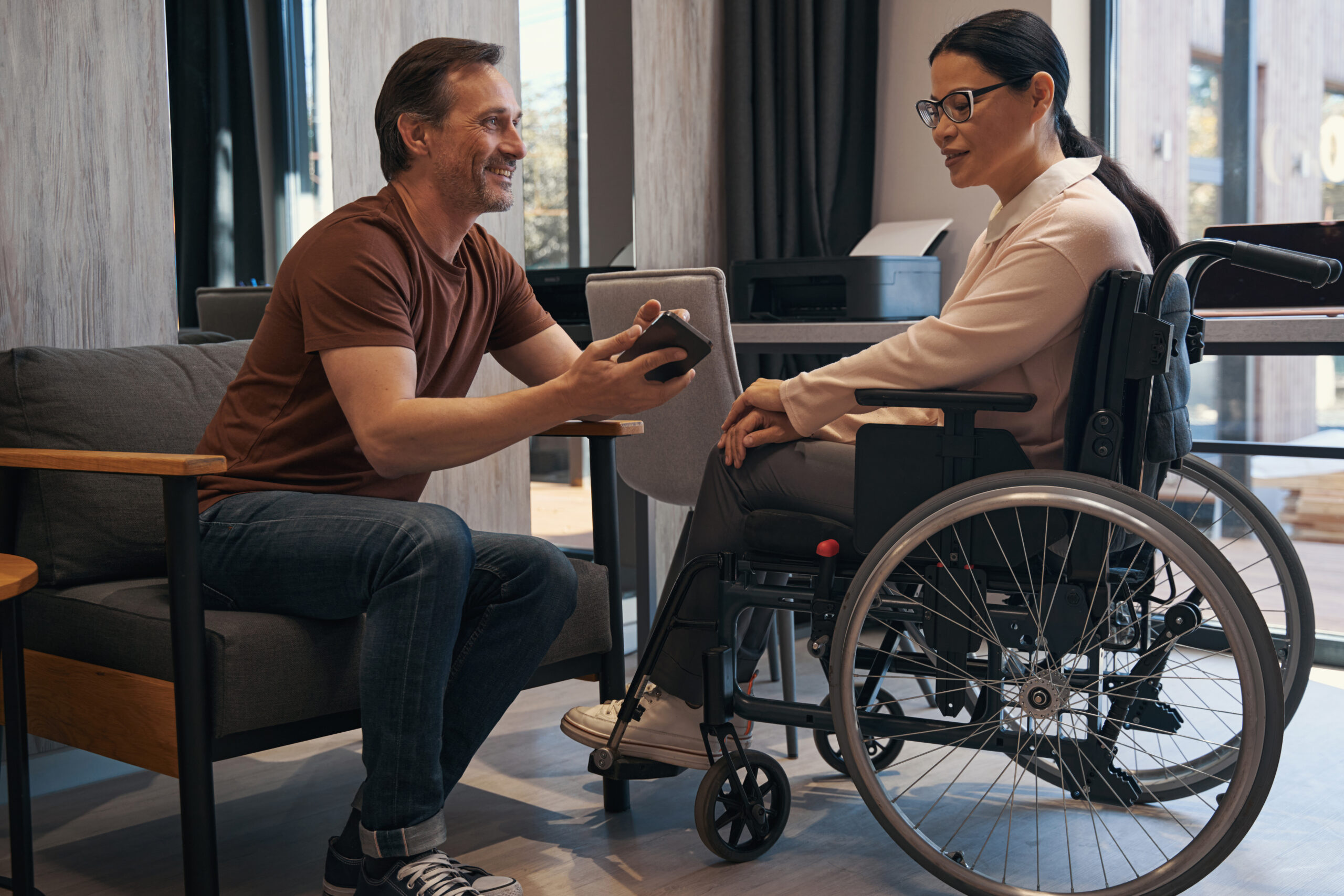 Cheerful mature male is showing his cell phone to elegant Asian woman using wheelchair indoors